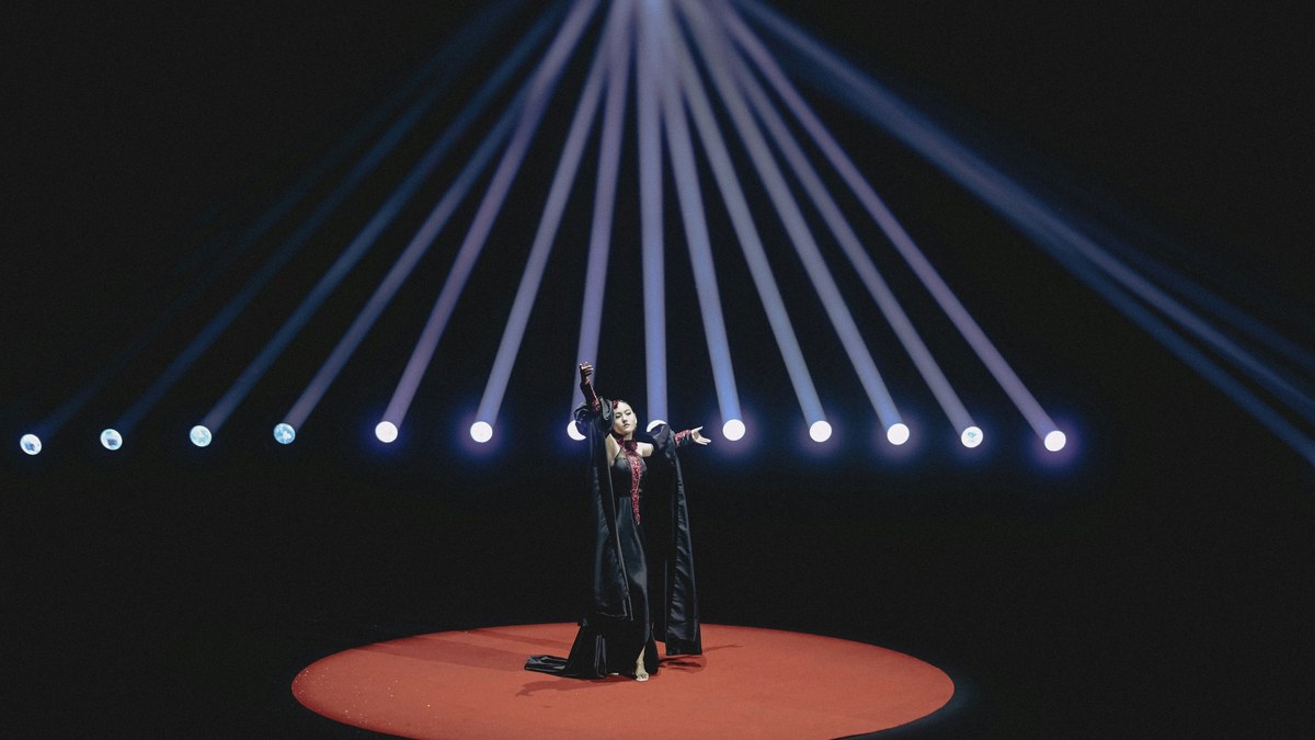 A solo performer on a red-lit round stage under a fan of light beams.