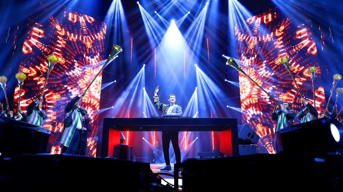 A DJ performing with horn players in front of red LED video walls.