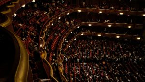A packed Broadway theatre auditorium with tiered red seats and balconies
