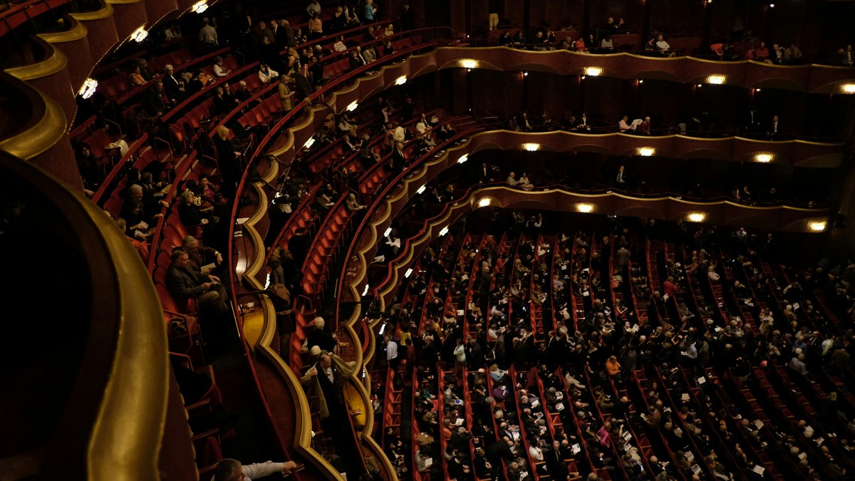 A packed Broadway theatre auditorium with tiered red seats and balconies