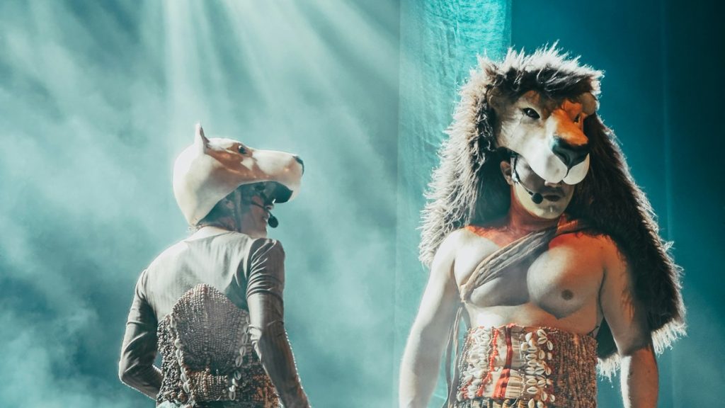 Two performers in animal masks on a smoke-filled stage under spotlights
