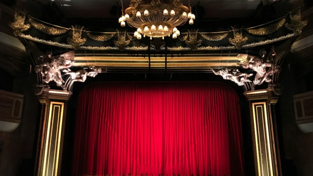 An ornate theatre proscenium with a red curtain and chandelier