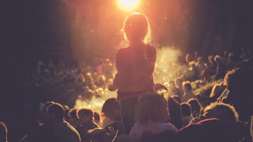 A child on a parent's shoulders watching an outdoor concert at sunset